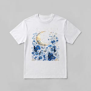 Op de afbeelding: Een wit T-shirt met korte mouwen en een centrale grafische print van een gouden halve maan, omringd door een veld met levendige blauwe bloemen, fijne stengels en kleine gouden steraccenten verspreid over het bovenste deel van het ontwerp.