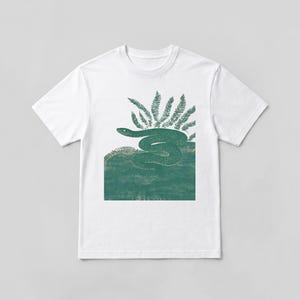May include: A white short-sleeved t-shirt featuring a central graphic print of a dark green snake coiled on a textured green surface, positioned below a cluster of dark green fern leaves. The design has a detailed, etched, or woodcut appearance.