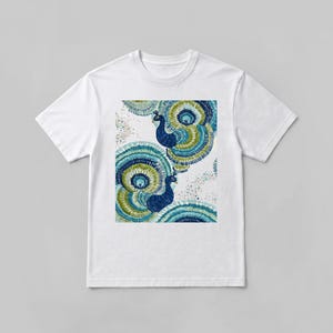 May include: A white short-sleeved t-shirt featuring a central graphic print of two peacocks with vibrant blue, green, and teal plumage. The design displays circular, fan-like tail feathers and speckled accents surrounding the birds on the fabric.