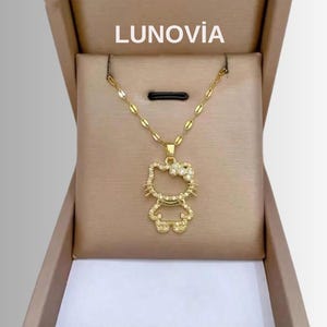 Puede incluir: Un collar dorado con un colgante con forma de gato de dibujos animados, incrustado con pequeñas piedras preciosas transparentes. El collar se muestra en una caja marrón con el nombre de la marca "LUNOVIA" impreso en la parte superior.