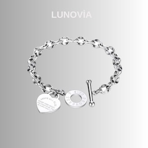 May include: A silver-colored bracelet with a chain-link design. The bracelet features a heart-shaped charm with engraved text and a circular charm with the text "PROVERBS 4:23". The brand name "LUNOVIA" is at the top.