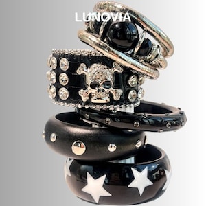 Puede incluir: Una pila de pulseras negras y plateadas. Las pulseras incluyen una cadena de serpiente plateada, una pulsera de cuentas negras, una pulsera de cuero negro con un diseño de calavera y huesos cruzados, una pulsera de madera negra con tachuelas plateadas y una pulsera negra con estrellas blancas.