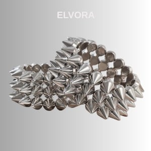 Può includere: Un braccialetto color argento con un motivo ripetuto di punte a forma di cono. Il braccialetto in metallo ha una finitura lucida. La parola "ELVORA" è stampata in bianco nella parte superiore dell'immagine.