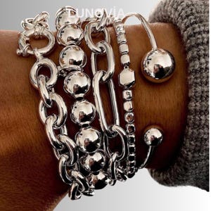 Puede incluir: Una colección de pulseras plateadas con varios diseños, incluyendo eslabones de cadena, hilos de cuentas y una barra curva con una esfera. Las pulseras se usan juntas, creando un aspecto en capas. Las pulseras son brillantes y reflectantes.
