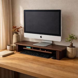 Peut inclure: Un support d'écran en bois brun foncé avec une étagère pour clavier, contenant un clavier et une souris noirs. Un ordinateur Apple argenté est posé dessus. Une petite plante en pot et un vase avec des fleurs séchées sont sur le bureau.