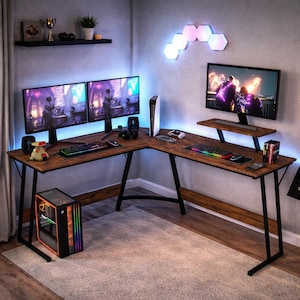 May include: A brown and black L-shaped gaming desk with multiple monitors, a gaming PC, and accessories. The desk has a wood-grain finish and black metal legs. The monitors display a video game scene. The room is lit with blue and pink LED lights.
