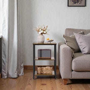 May include: A three-tiered side table with a dark brown top and shelves, supported by a black metal frame. The top shelf holds a vase of dried flowers and a small bowl. A speaker and a woven basket are on the lower shelves.