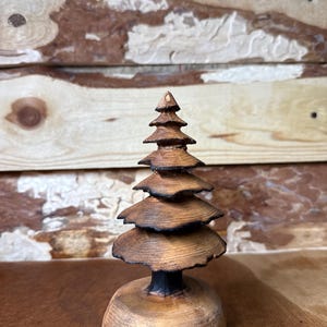 May include: A handcrafted wooden Christmas tree sculpture. The tree features layered, circular tiers, with a dark, charred trunk and base. The natural wood grain is visible, and the sculpture is set against a rustic wooden backdrop.