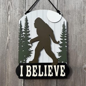 May include: Wooden sign depicting a Bigfoot silhouette walking past pine trees and a moon. The sign is white with a black border and the words "I BELIEVE" in white.