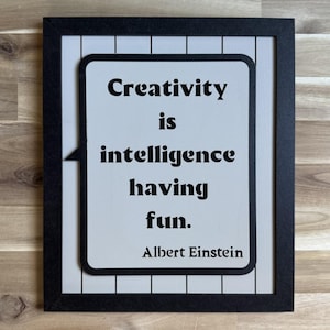 May include: A framed sign with the quote "Creativity is intelligence having fun." in black text on a light gray background. The sign is in a black frame and is attributed to Albert Einstein.