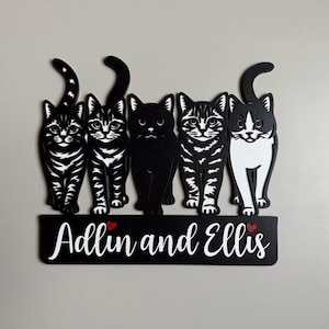 May include: A black and white wooden sign featuring five stylized cat figures. The cats are in various poses, with a mix of black, white, and tabby patterns. Below the cats is a black banner with the names "Adllin and Ellis" in white script, with red hearts.