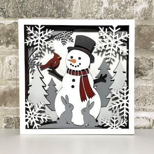 May include: A layered, framed winter scene features a snowman with a top hat and scarf, a red cardinal, two gray rabbits, and stylized trees. White snowflakes frame the scene. The artwork is set against a brick background.