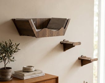May include: A wall-mounted dark wood cat bed with a gray cushioned interior and two matching dark wood wall-mounted steps. The bed features a curved front edge and is secured with visible metal screws, providing a elevated resting area.