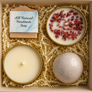 May include: A gift box containing an "All Natural Handmade Soap" bar, two candles in gold tins, one with red petals, and a white bath bomb. The items are nestled in shredded paper.