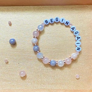 May include: A circular beaded bracelet featuring white, light pink, and grey beads with black letter beads spelling out the phrase BEGIN AGAIN. The bracelet is placed inside a rectangular gold-colored wooden tray with side handles.