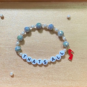 May include: A beaded bracelet featuring white round beads with black letters spelling out "THATS ALL," interspersed with small white pearls, larger iridescent blue-grey beads, and a single red high-heeled shoe charm attached to the side.