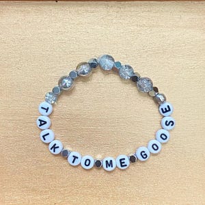 May include: A beaded bracelet featuring white circular beads with black lettering that spells out TALK TO ME GOOSE. The bracelet is constructed with a mix of clear, textured, and metallic-toned beads interspersed between the lettered beads.