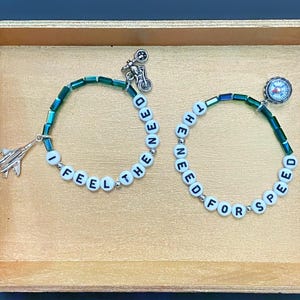 May include: Two beaded bracelets featuring white letter beads spelling I FEEL THE NEED and THE NEED FOR SPEED. The bracelets include green cylindrical beads and silver-toned charms shaped like fighter jets, a motorcycle, and a small compass.