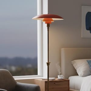 Puede incluir: Una l&aacute;mpara de pie moderna con una pantalla escalonada de color naranja rojizo y un tallo color lat&oacute;n. La l&aacute;mpara se apoya sobre una base circular negra. La l&aacute;mpara est&aacute; en un dormitorio, junto a una ventana y una mesita de noche de madera.