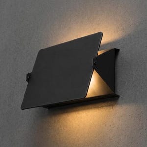 Könnte beinhalten: Eine mattschwarze Wandleuchte mit einer rechteckigen Metallplatte, die eine Lichtquelle teilweise verdeckt und einen warmen, goldenen Schein sowohl oberhalb als auch unterhalb der Leuchte auf eine strukturierte graue Fläche wirft.