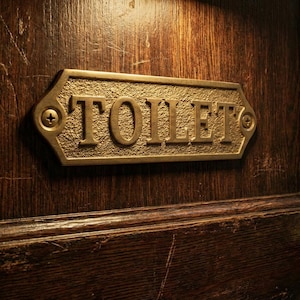 Könnte beinhalten: Ein rechteckiges Messingschild mit dem eingeprägten Wort TOILET in Großbuchstaben, das mit zwei Schrauben an einer dunkelbraunen Holztür befestigt ist. Das Schild hat einen strukturierten Hintergrund und einen glatten Rand über einem runden Türknauf.