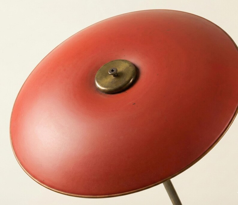 Puede incluir: Una pantalla de l&aacute;mpara roja de forma ovalada con un accesorio central de color lat&oacute;n. La pantalla tiene un acabado liso y mate y un fino borde de color lat&oacute;n alrededor del borde. El tallo de la l&aacute;mpara es visible en la parte inferior.
