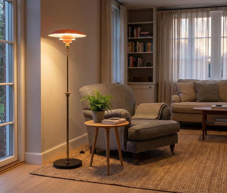 Puede incluir: Una l&aacute;mpara de pie de estilo retro con una pantalla escalonada de color naranja-rojo y un poste de color lat&oacute;n. La l&aacute;mpara est&aacute; colocada junto a un sill&oacute;n gris y una peque&ntilde;a mesa auxiliar de madera con una planta en maceta. La habitaci&oacute;n tiene una paleta de colores neutros con una alfombra.