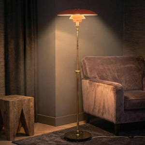 Puede incluir: Una l&aacute;mpara de pie de mediados de siglo con una pantalla escalonada de color naranja rojizo y un tallo color lat&oacute;n. La l&aacute;mpara est&aacute; colocada en una habitaci&oacute;n con un sill&oacute;n marr&oacute;n oscuro y una mesa auxiliar de madera. El dise&ntilde;o de la l&aacute;mpara es un cl&aacute;sico.