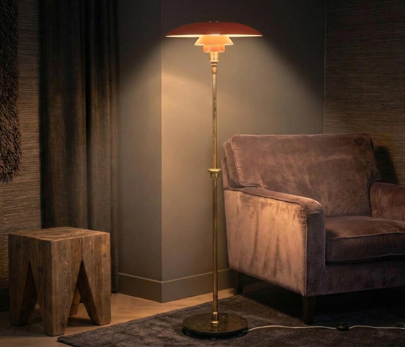 Puede incluir: Una l&aacute;mpara de pie de mediados de siglo con una pantalla escalonada de color naranja rojizo y un tallo color lat&oacute;n. La l&aacute;mpara est&aacute; colocada en una habitaci&oacute;n con un sill&oacute;n marr&oacute;n oscuro y una mesa auxiliar de madera. El dise&ntilde;o de la l&aacute;mpara es un cl&aacute;sico.