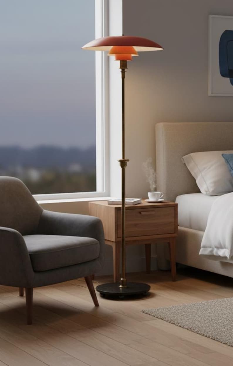 Puede incluir: Una l&aacute;mpara de pie moderna con una pantalla escalonada de color naranja rojizo y un tallo color lat&oacute;n. La l&aacute;mpara se apoya sobre una base circular negra. La l&aacute;mpara est&aacute; en un dormitorio, junto a una ventana y una mesita de noche de madera.