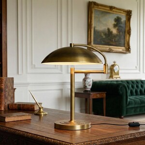 Pode incluir: Uma lumin&aacute;ria de mesa de lat&atilde;o com uma c&uacute;pula e bra&ccedil;o ajust&aacute;vel est&aacute; em uma mesa de madeira. A lumin&aacute;ria est&aacute; acesa, emitindo um brilho quente. A mesa est&aacute; em uma sala com uma estante, livros e um sof&aacute; verde.
