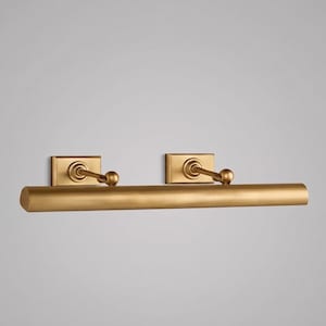 May include: A gold-colored picture light fixture with two rectangular wall mounts and a horizontal cylindrical light bar. Each mount has a curved arm and a small ball at the end. The fixture is designed for illuminating artwork.