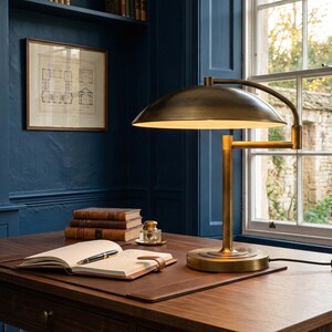 Pode incluir: Uma lumin&aacute;ria de mesa de lat&atilde;o com um bra&ccedil;o curvo e uma c&uacute;pula est&aacute; em uma mesa de madeira. Um caderno de couro com uma caneta, uma pilha de livros e um desenho arquitet&ocirc;nico emoldurado tamb&eacute;m est&atilde;o na mesa. O fundo apresenta uma parede azul e uma janela.