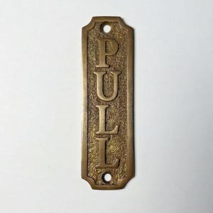 May include: A rectangular brass door plate with a textured background and the word PULL embossed in capital letters. The plate features rounded corners and two circular screw holes, one at the top and one at the bottom, for mounting purposes.