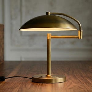 Pode incluir: Uma lumin&aacute;ria de mesa de lat&atilde;o com um bra&ccedil;o curvo e uma c&uacute;pula. A lumin&aacute;ria tem uma base redonda e um suporte vertical. A lumin&aacute;ria est&aacute; em uma superf&iacute;cie de madeira. A lumin&aacute;ria est&aacute; ligada.