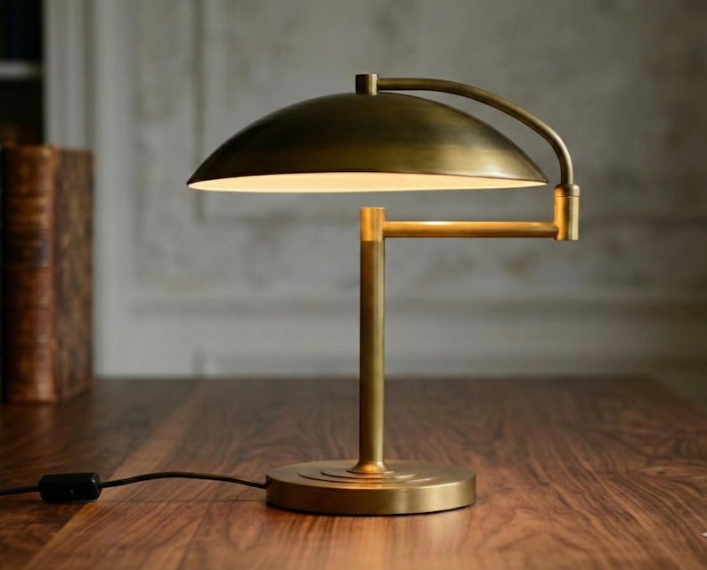 Pode incluir: Uma lumin&aacute;ria de mesa de lat&atilde;o com um bra&ccedil;o curvo e uma c&uacute;pula. A lumin&aacute;ria tem uma base redonda e um suporte vertical. A lumin&aacute;ria est&aacute; em uma superf&iacute;cie de madeira. A lumin&aacute;ria est&aacute; ligada.