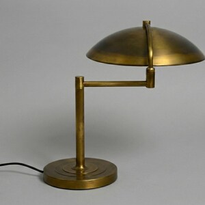Pode incluir: Uma lumin&aacute;ria de mesa em tom de lat&atilde;o com uma c&uacute;pula e um bra&ccedil;o ajust&aacute;vel. A l&acirc;mpada tem uma base redonda e um cabo de alimenta&ccedil;&atilde;o preto. O design lembra a ilumina&ccedil;&atilde;o industrial vintage.