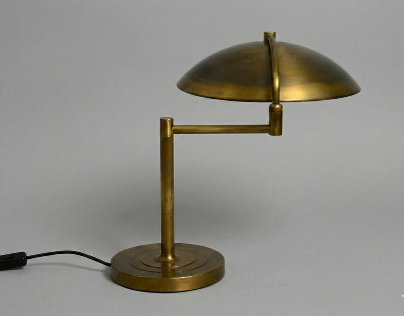 Pode incluir: Uma lumin&aacute;ria de mesa em tom de lat&atilde;o com uma c&uacute;pula e um bra&ccedil;o ajust&aacute;vel. A l&acirc;mpada tem uma base redonda e um cabo de alimenta&ccedil;&atilde;o preto. O design lembra a ilumina&ccedil;&atilde;o industrial vintage.