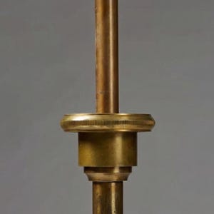 Puede incluir: Primer plano de un componente de l&aacute;mpara de lat&oacute;n. El dise&ntilde;o presenta un v&aacute;stago cil&iacute;ndrico con un anillo decorativo estriado y una serie de elementos redondeados apilados. El lat&oacute;n tiene una p&aacute;tina c&aacute;lida y envejecida, lo que sugiere or&iacute;genes vintage o antiguos.