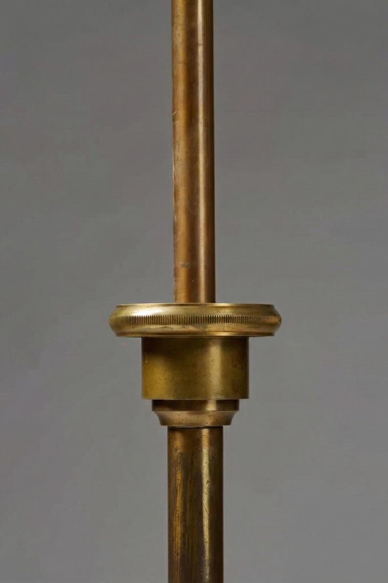 Puede incluir: Primer plano de un componente de l&aacute;mpara de lat&oacute;n. El dise&ntilde;o presenta un v&aacute;stago cil&iacute;ndrico con un anillo decorativo estriado y una serie de elementos redondeados apilados. El lat&oacute;n tiene una p&aacute;tina c&aacute;lida y envejecida, lo que sugiere or&iacute;genes vintage o antiguos.
