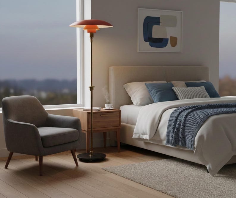 Puede incluir: Un dormitorio moderno con una l&aacute;mpara de pie con pantalla roja, un sill&oacute;n gris y una cama con almohadas y una manta azules. La l&aacute;mpara tiene un poste de lat&oacute;n y una base redonda. Una mesita de noche de madera y una obra de arte abstracta enmarcada completan la decoraci&oacute;n.