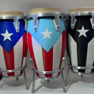 Könnte beinhalten: Drei Conga-Trommeln mit dem Design der puerto-ricanischen Flagge. Die Trommeln sind blau, hellblau und schwarz, mit weißen Sternen und rot-weißen Streifen. Jede Trommel hat einen silbernen Rand und steht auf einem silbernen Ständer.