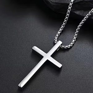 Op de afbeelding: Een zilverkleurige kruisvormige hangerketting. Het kruis heeft een eenvoudig, gepolijst ontwerp met een glad oppervlak. De ketting is een schakelstijl, eveneens zilverkleurig, en de achtergrond is donker.