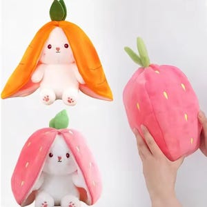 Könnte beinhalten: Drei Plüschtiere: ein weißes Kaninchen in einem orangefarbenen Karottenkostüm, ein weißes Kaninchen in einem rosa Erdbeerkostüm und ein rosa Erdbeerplüsch. Die Spielzeuge haben gestickte Gesichtsmerkmale und sind aus weichem Stoff.