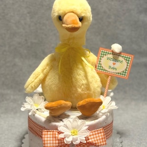 May include: A yellow plush duckling with an orange beak and feet sits atop a diaper cake. The cake is decorated with white daisies and an orange and white gingham ribbon. A sign that says "Welcome Baby" is held by the duckling.