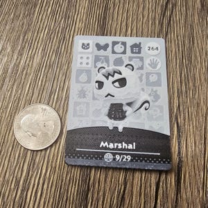 May include: A gray and white trading card featuring the character Marshal from Animal Crossing. The card has a black border and includes the number 264. A U.S. quarter is next to the card.