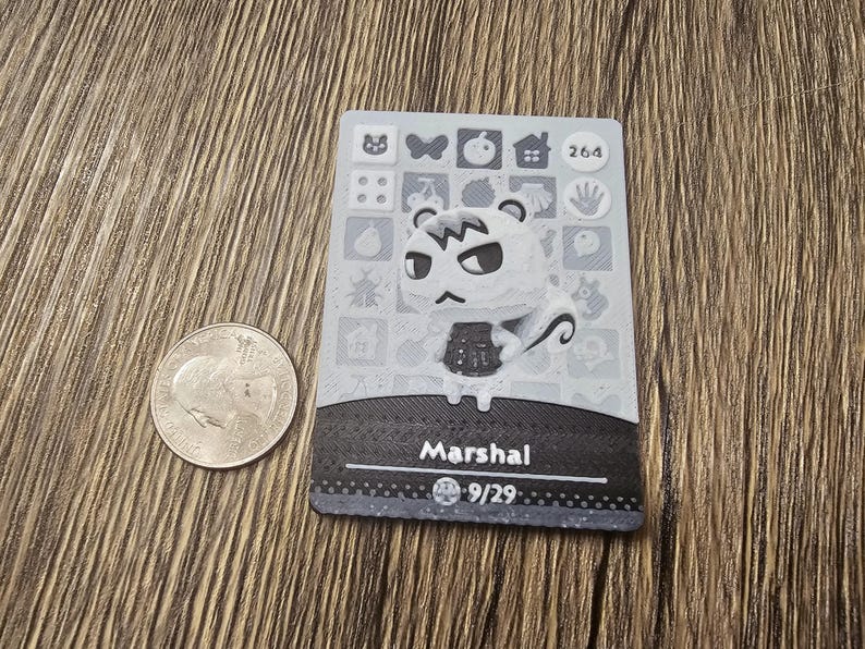 May include: A gray and white trading card featuring the character Marshal from Animal Crossing. The card has a black border and includes the number 264. A U.S. quarter is next to the card.