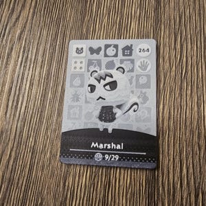 May include: A grey and white Animal Crossing trading card featuring Marshal. The card displays an illustration of Marshal, his name, and a number. The rectangular card has a patterned border and is set against a wooden background.