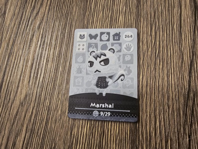 May include: A grey and white Animal Crossing trading card featuring Marshal. The card displays an illustration of Marshal, his name, and a number. The rectangular card has a patterned border and is set against a wooden background.