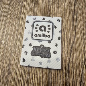 May include: Amiibo card featuring the Animal Crossing logo. The card has a white background with a pattern of small black icons. The Amiibo logo is in a white square with a black border. The Animal Crossing logo is in a black banner.